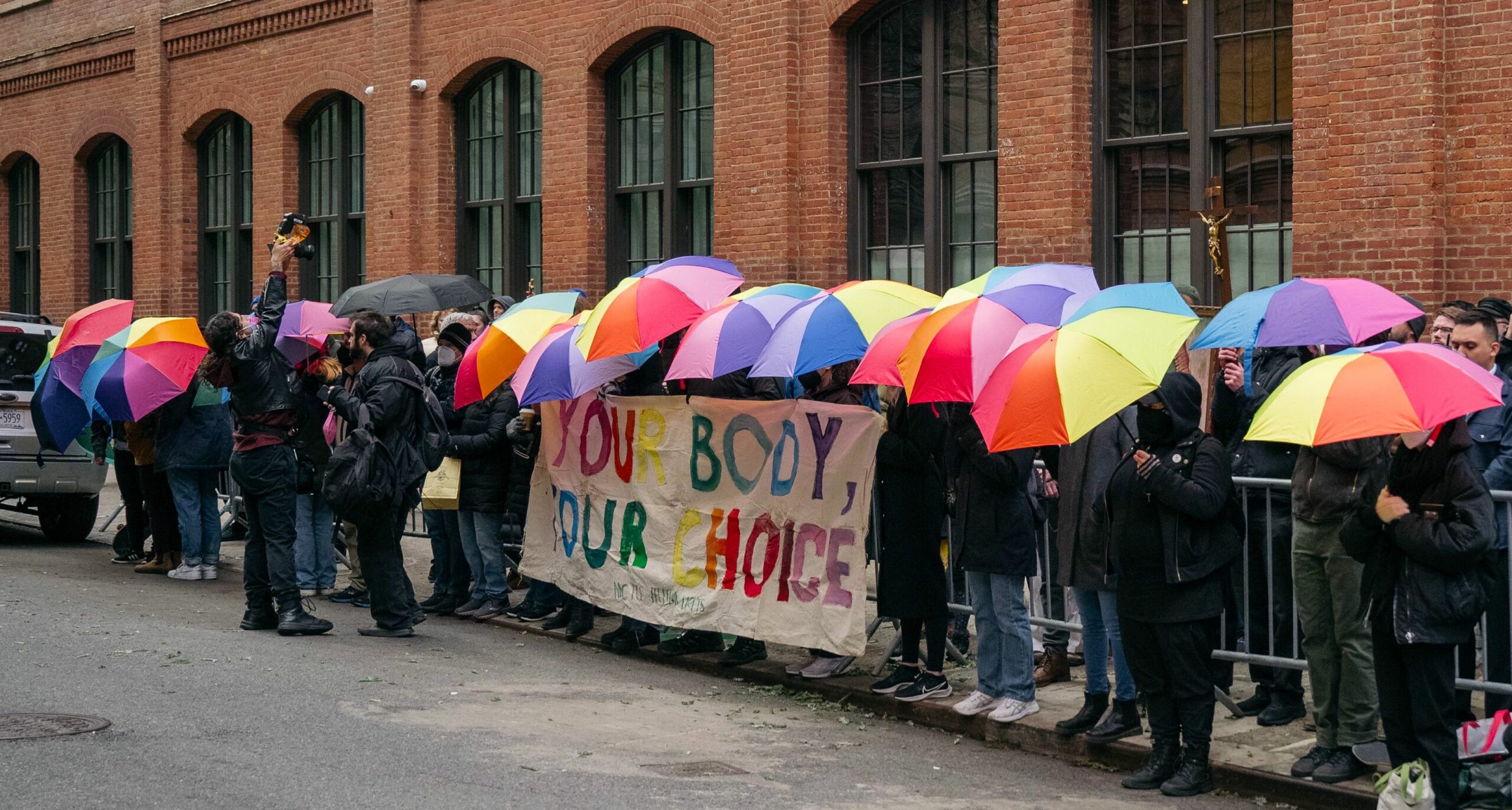 Clinic defenders block anti-abortion harassers with rainbow umbrellas and a big banner that reads "Your Body Your Choice"